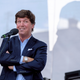 Tucker Carlson stands at a microphone with is arms crossed.
